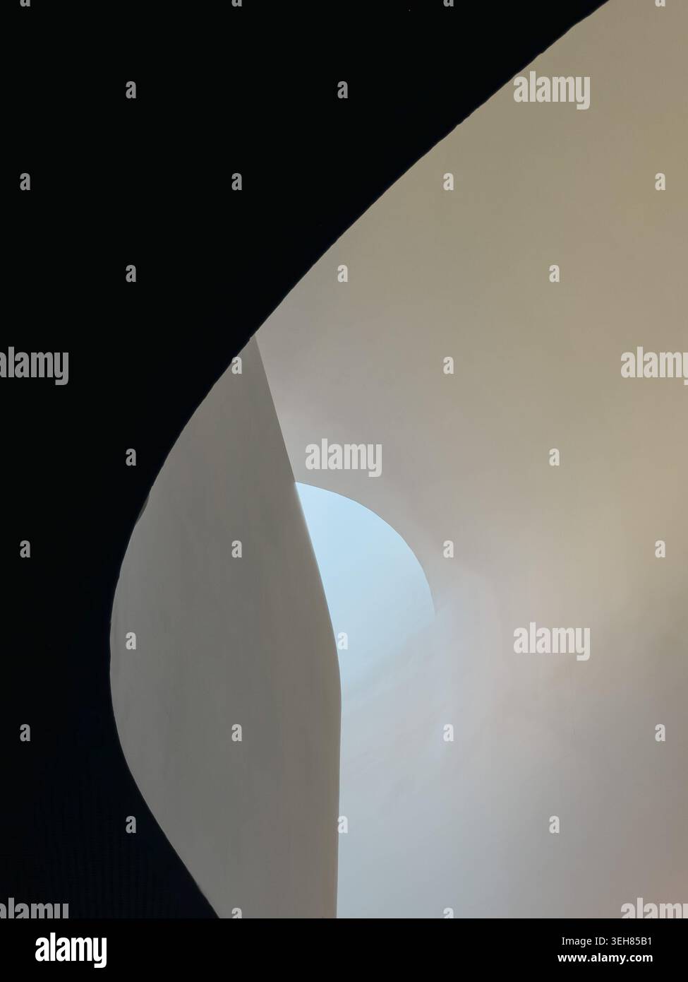 Una superficie architettonica bianca curva e liscia si estende lungo la cornice con una piccola apertura ad arco che rivela un cielo azzurro pallido, adagiato su un'ombra profonda in un'audace composizione architettonica minimalista. Foto Stock