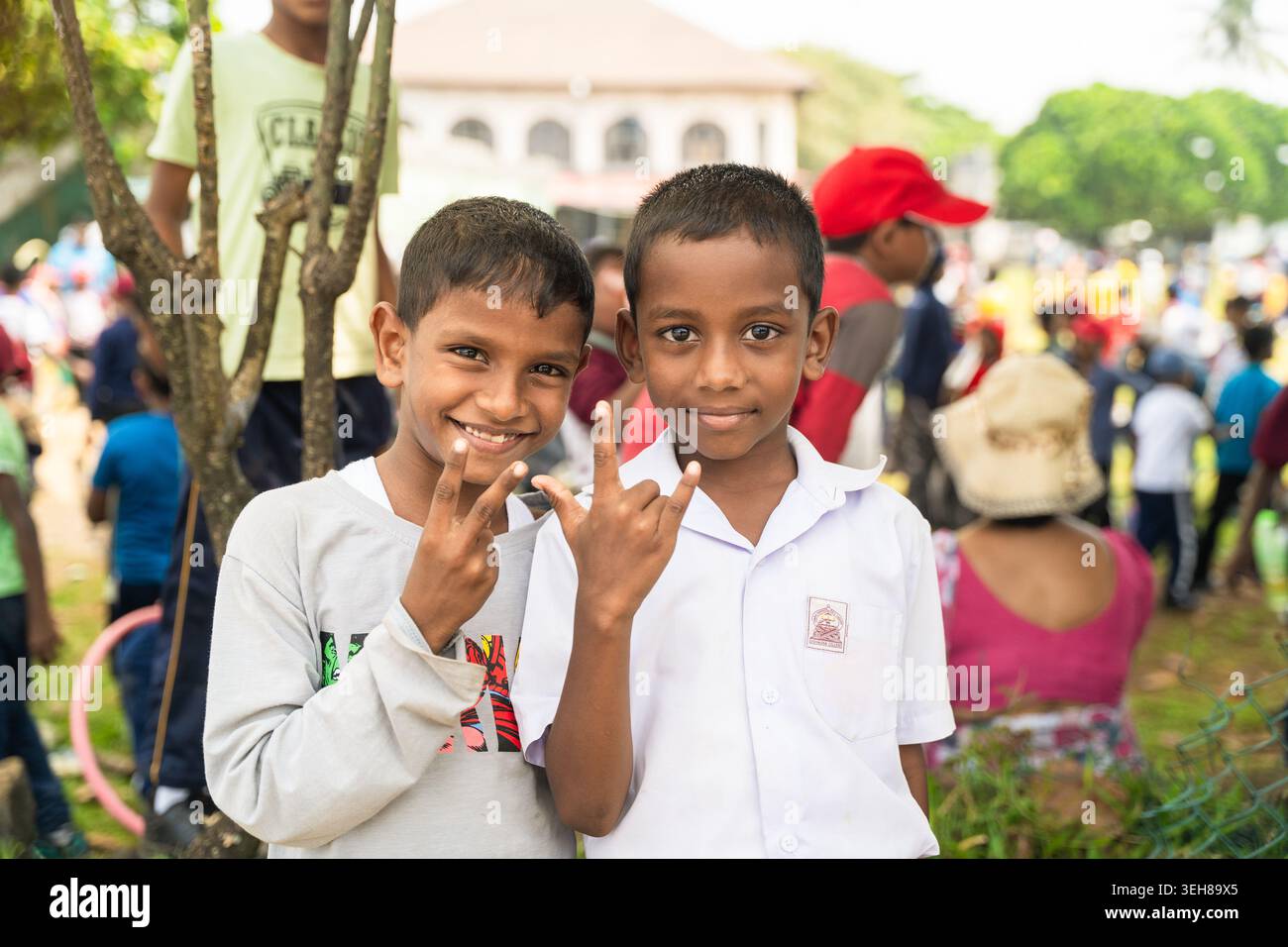 Sri Lanka - 12 febbraio 2026: Vista di due studenti sorridenti che fanno gesti manuali durante un evento all'aperto con una folla e una vegetazione sullo sfondo. Foto Stock