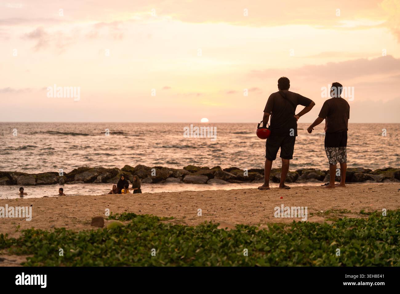 Sri Lanka - 14 gennaio 2026: Vista di due uomini in piedi su una spiaggia sabbiosa che guardano il tramonto sull'Oceano Indiano con persone che nuotano vicino alla riva. Foto Stock