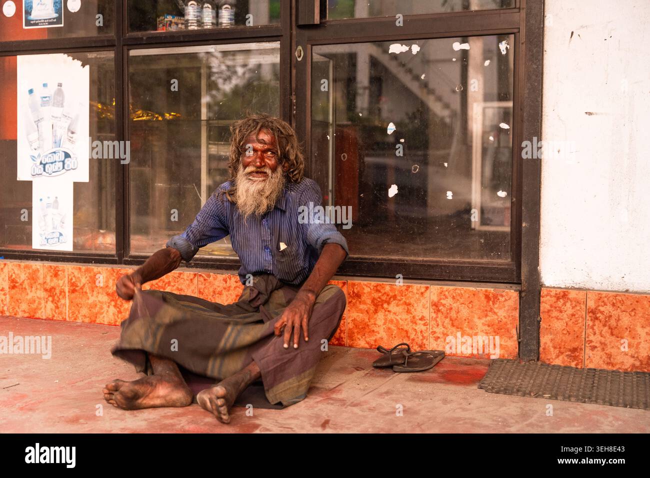 Sri Lanka - 14 gennaio 2026: Vista di un uomo anziano con una barba lunga e capelli disordinati seduto a terra di fronte a una vetrina con poster. Foto Stock
