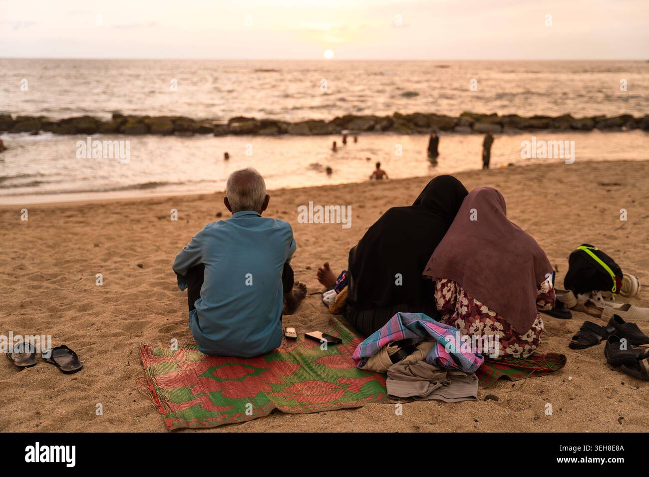 Sri Lanka - 14 gennaio 2026: Veduta di un uomo e di due donne seduti sulla sabbia che guardano il tramonto sull'Oceano Indiano con onde dolci e un morbido sk Foto Stock