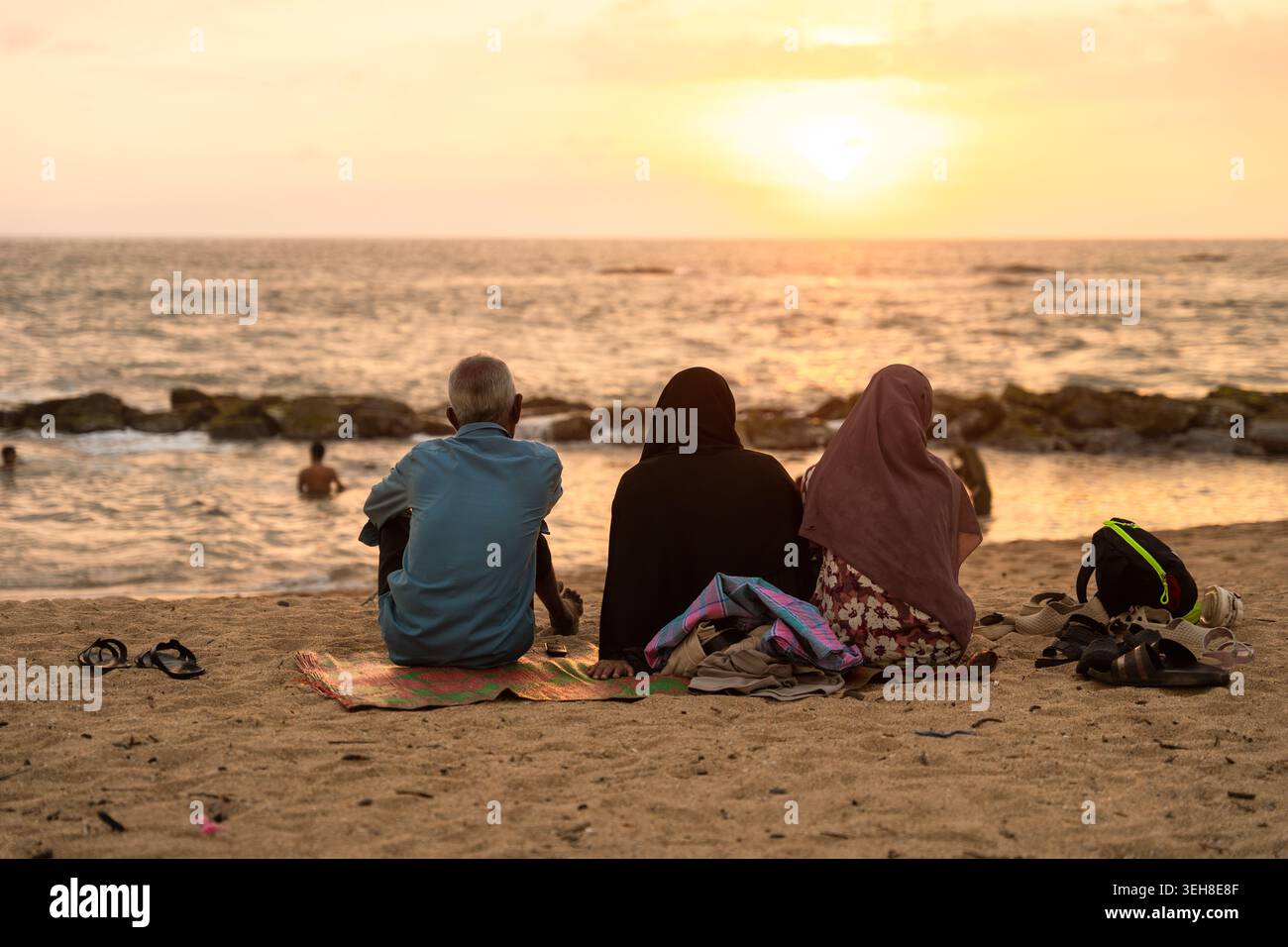 Sri Lanka - 14 gennaio 2026: Veduta di tre persone sedute su una spiaggia sabbiosa che guarda il tramonto sull'Oceano Indiano con frangiflutti rocciosi e nuotate Foto Stock