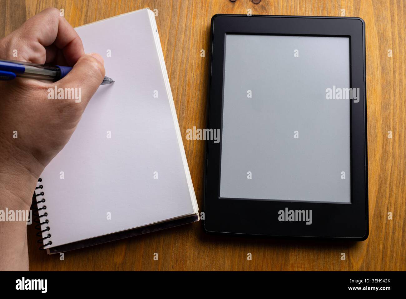 Una persona mancina scrive appunti in un quaderno mentre utilizza un lettore elettronico, seduto a un tavolo di legno duro Foto Stock
