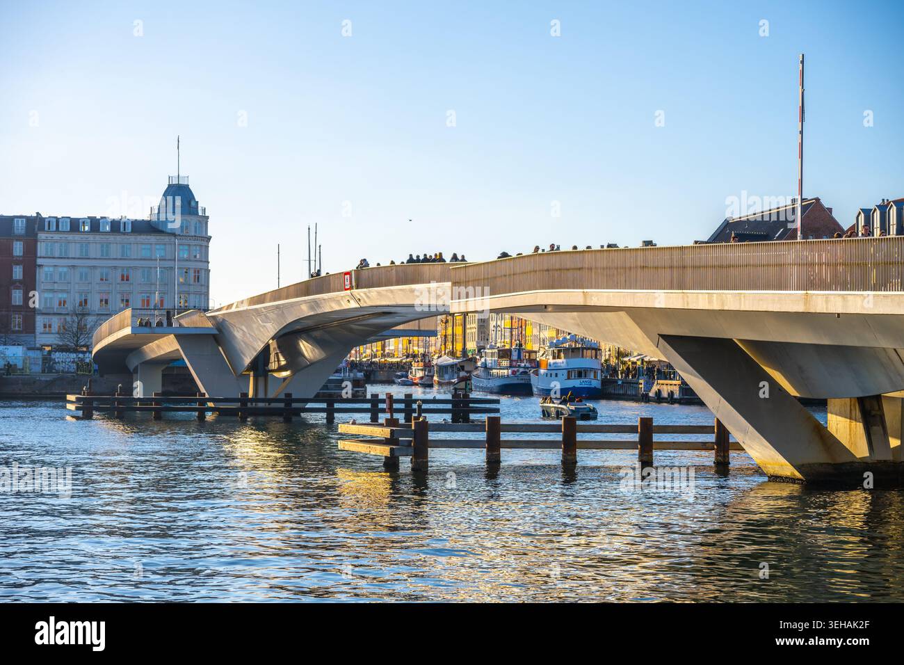 I visitatori attraversano a piedi l'Inner Harbour Bridge di Copenhagen, Danimarca. Il sole splende sull'acqua e sugli edifici mentre le barche galleggiano nel porto. Cavolo Foto Stock