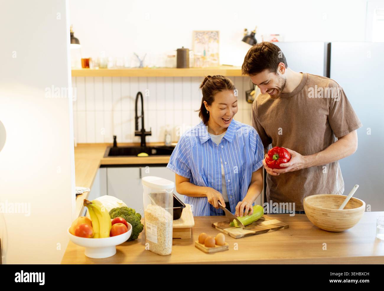 Una donna coreana e un uomo caucasico cucinano in una cucina moderna. Sta tritando verdure mentre lui tiene un pepe rosso. Sembrano felici ed enga Foto Stock