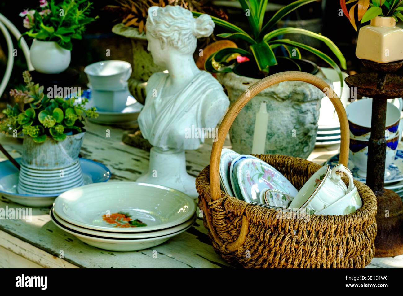 Un'affascinante esposizione all'aperto di antiche ceramiche, un busto femminile classico bianco e un cestino intrecciato pieno di tazze da tè vintage Foto Stock
