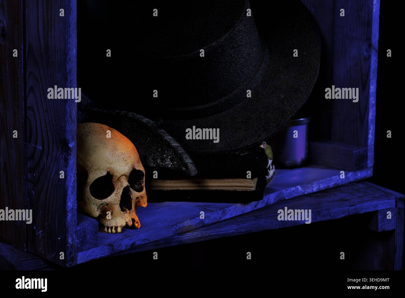 Un inquietante accordo di natura morta con un teschio, un cappello e un libro scuro sullo sfondo blu per le vibrazioni di Halloween Foto Stock