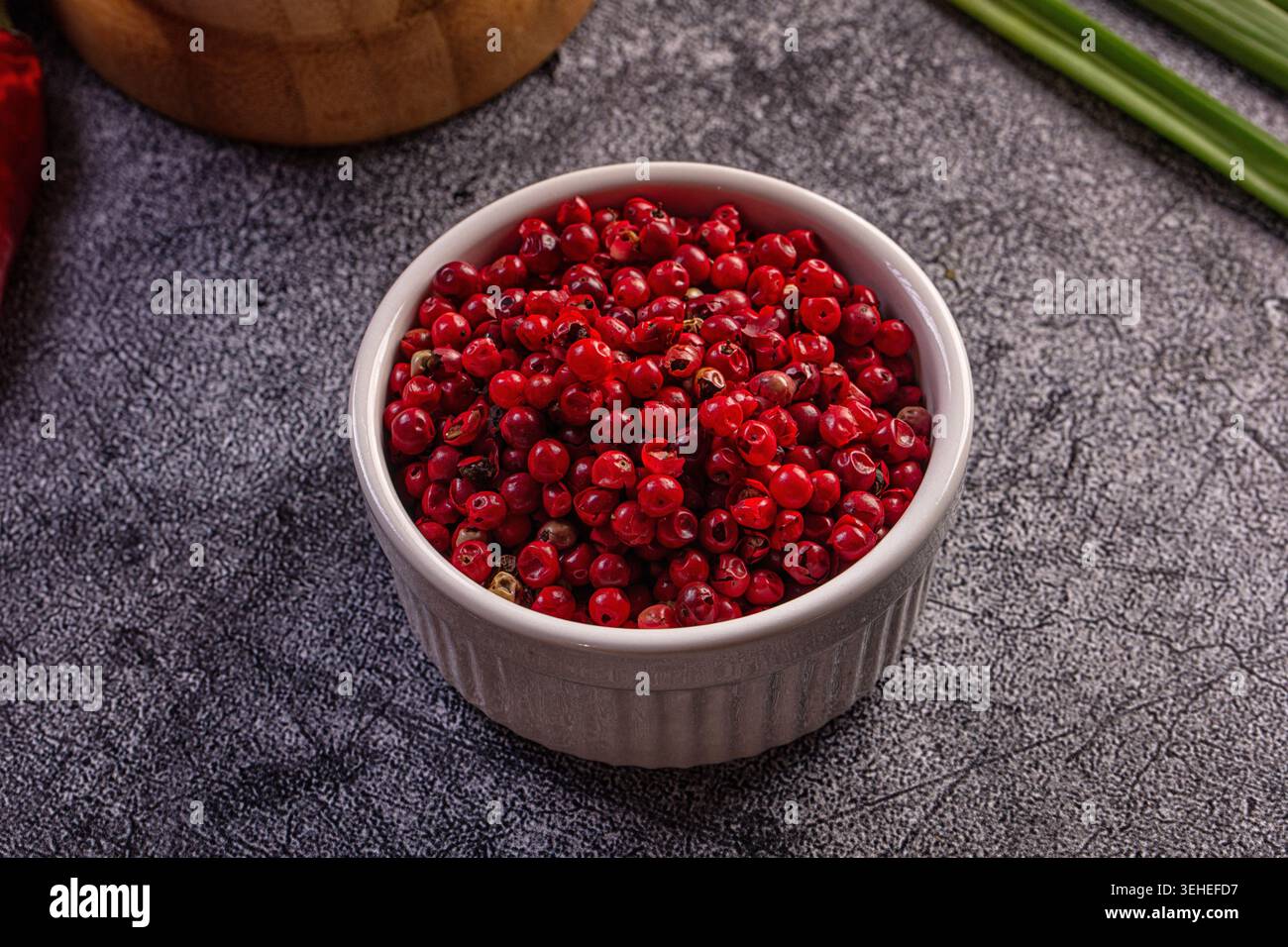 Primo piano di grani di pepe rosa rosso brillante in un recipiente di ceramica bianco. Sfondo rustico scuro con peperoncino rosso sfocato, mortaio di legno, cipolla verde Foto Stock
