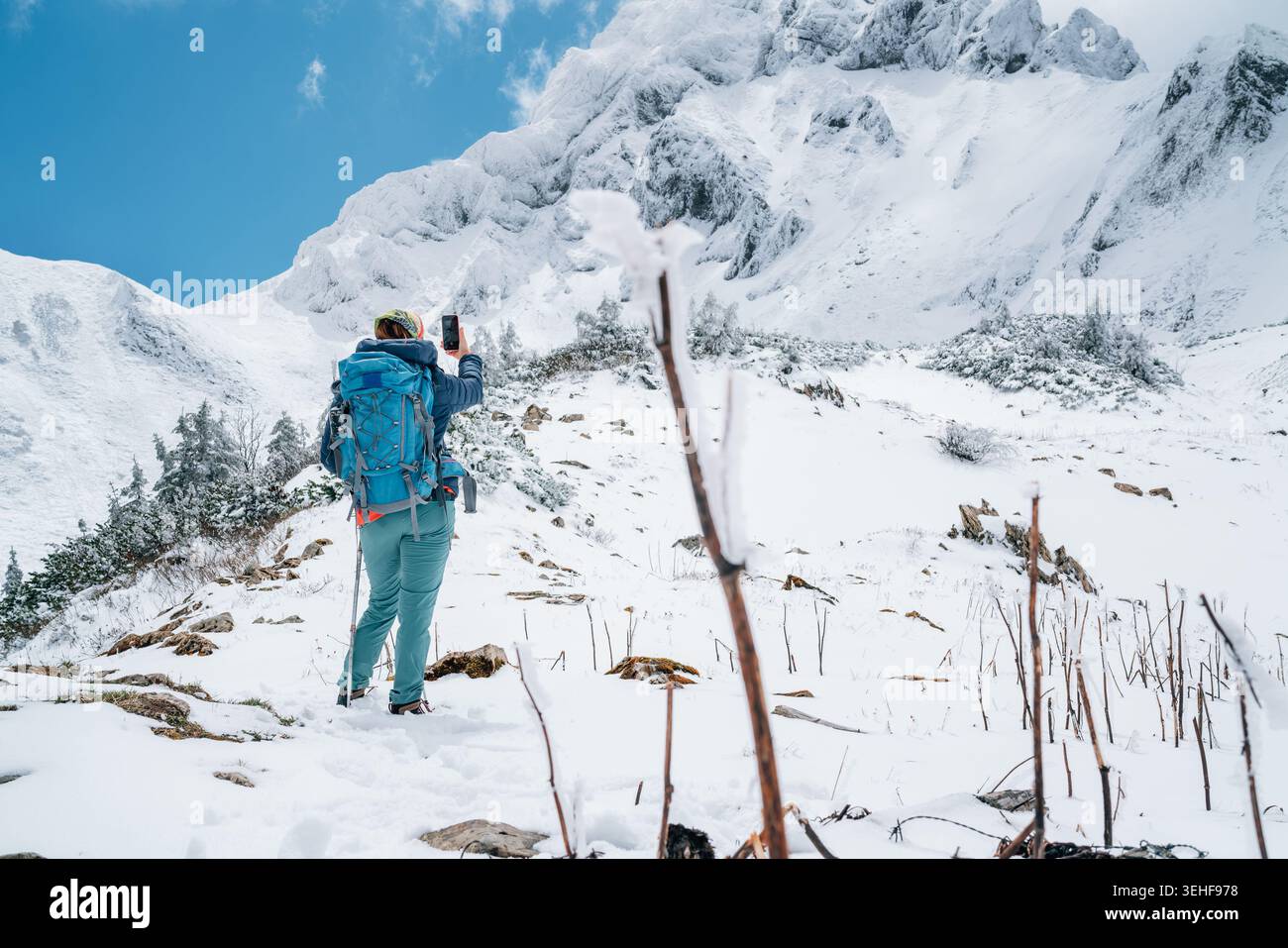 Trekker donna con zaino blu che scatta foto con lo smartphone della maestosa montagna Eisenerzer Reichenstein in Austria, che rappresenta l'avventura invernale Foto Stock