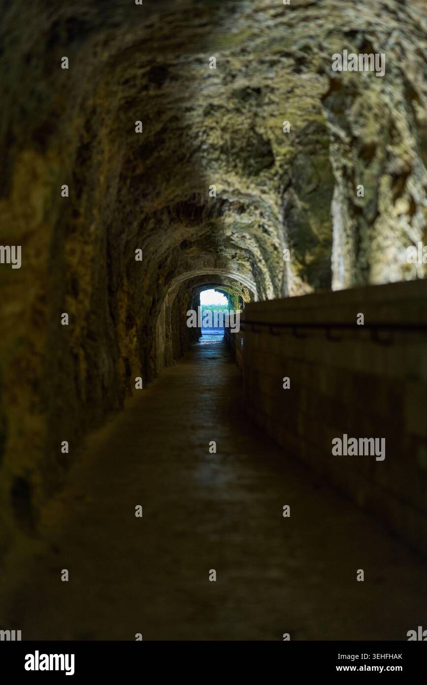 Tunnel sotterraneo lungo e stretto con aspre pareti in pietra e un passaggio pedonale che conduce in lontananza a un'uscita luminosa Foto Stock