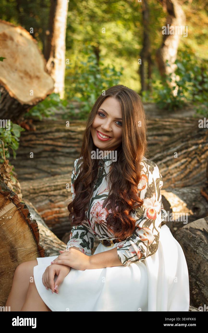 Una giovane donna con lunghi capelli castani siede su un tronco in una foresta. Indossa una blusa floreale e una gonna bianca, sorridendo in un ambiente naturale Foto Stock