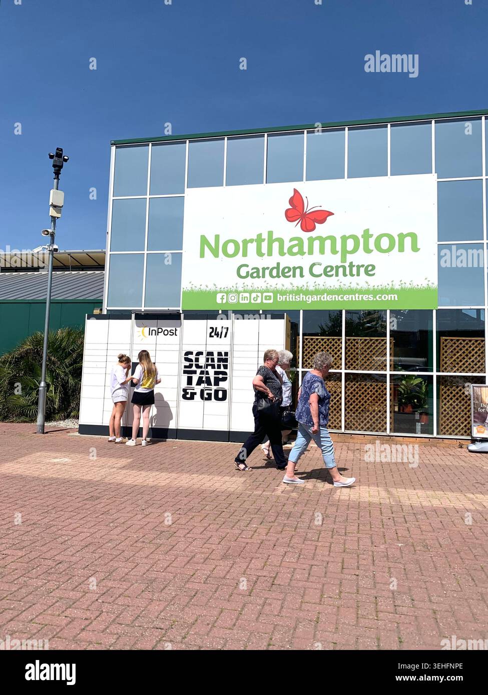 I visitatori passano davanti all'ingresso del Northampton Garden Centre Northamptonshire Inghilterra in un pomeriggio di sole con il logo della farfalla sulla facciata - Immagine stock catturata con smartphone