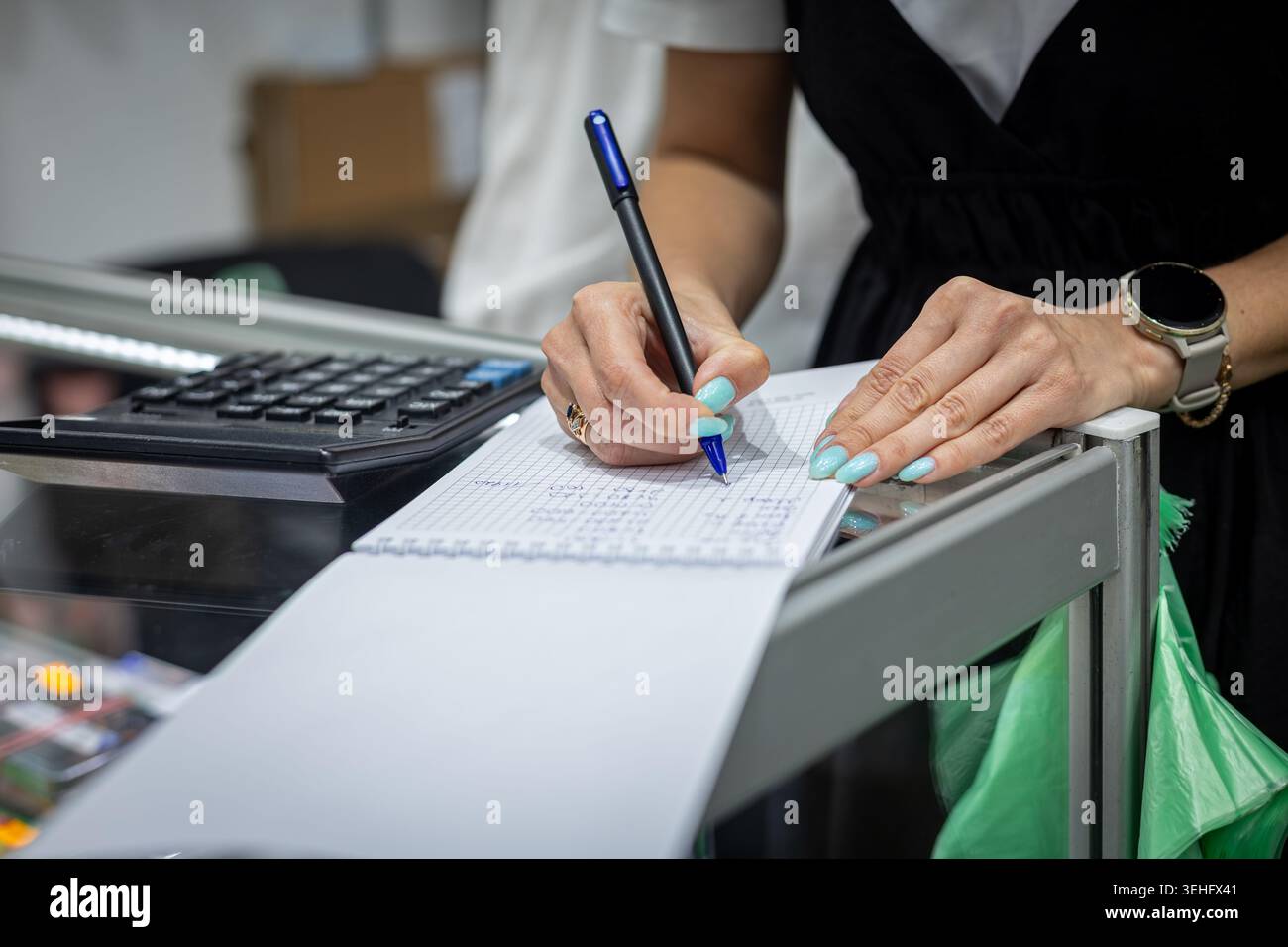 Il cassiere scrive la ricevuta al banco cassa. Primo piano di un lavoratore che scrive appunti su carta accanto a un registratore di cassa presso un banco cassa. Foto Stock