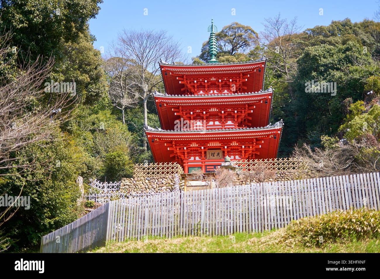 Una tradizionale pagoda giapponese a tre piani (sanju-no-to) con un'architettura in legno rosso vivace si erge in modo prominente contro uno sfondo di verde lussureggiante Foto Stock
