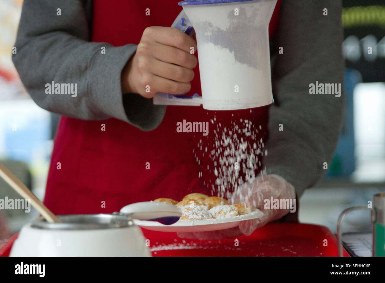Vista ravvicinata di un venditore in grembiule rosso che utilizza uno shaker setacciatore di farina di plastica per polverizzare lo zucchero a velo in polvere sui pancake caldi di poffertjes olandesi. Foto Stock