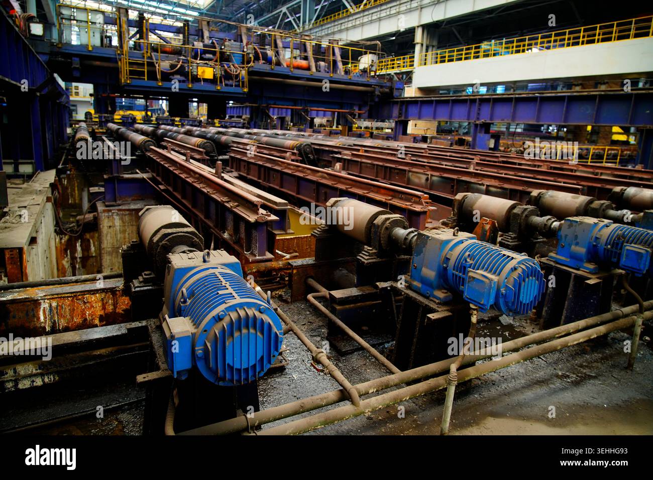 L'area di produzione principale di laminatoio a caldo, l'array di motori di azionamento blu e la tavola dei rulli di laminazione arrugginiti costituiscono l'officina industriale di formalina in acciaio Foto Stock