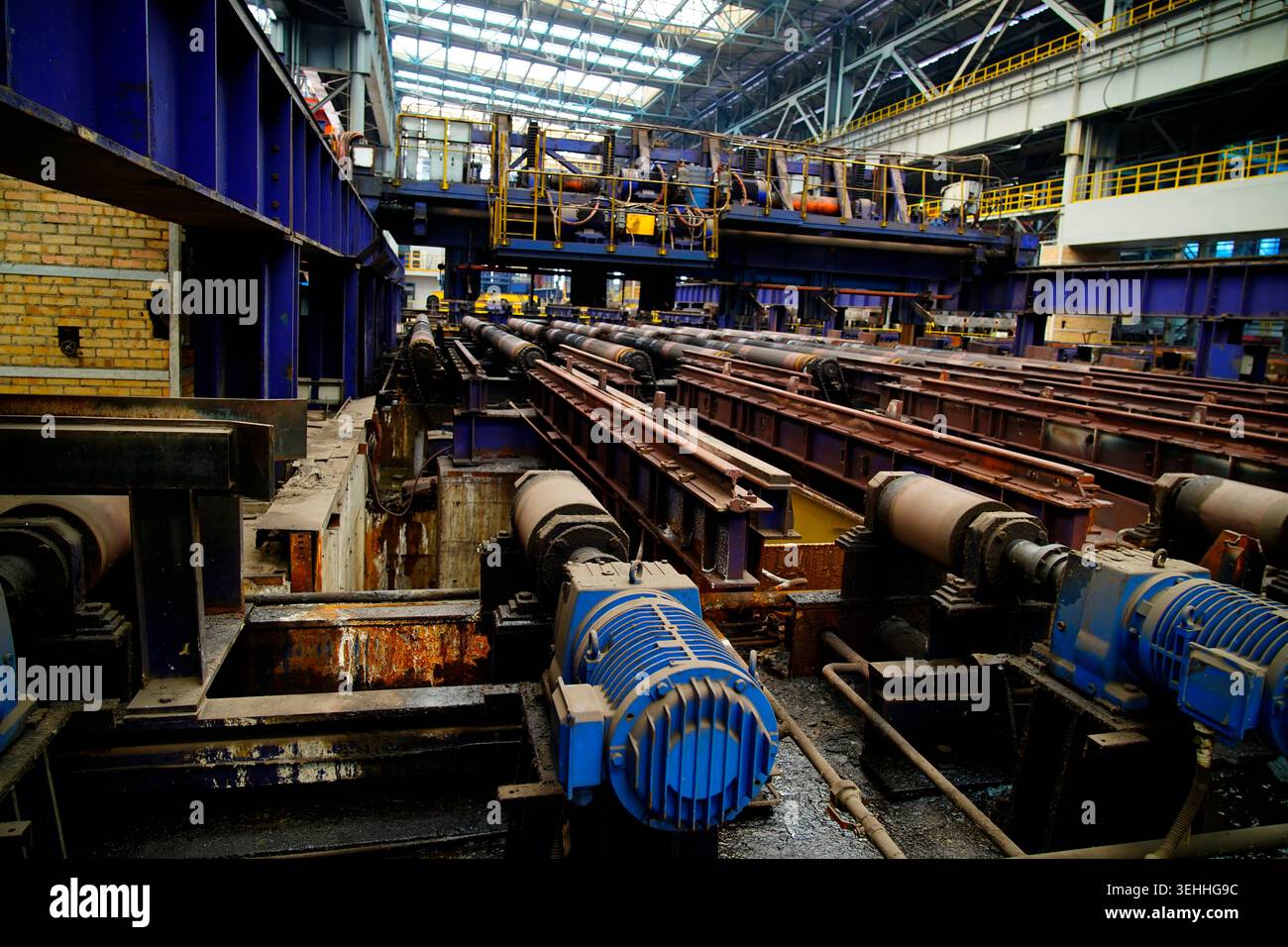 L'area di produzione principale di laminatoio a caldo, l'array di motori di azionamento blu e la tavola dei rulli di laminazione arrugginiti costituiscono l'officina industriale di formalina in acciaio Foto Stock