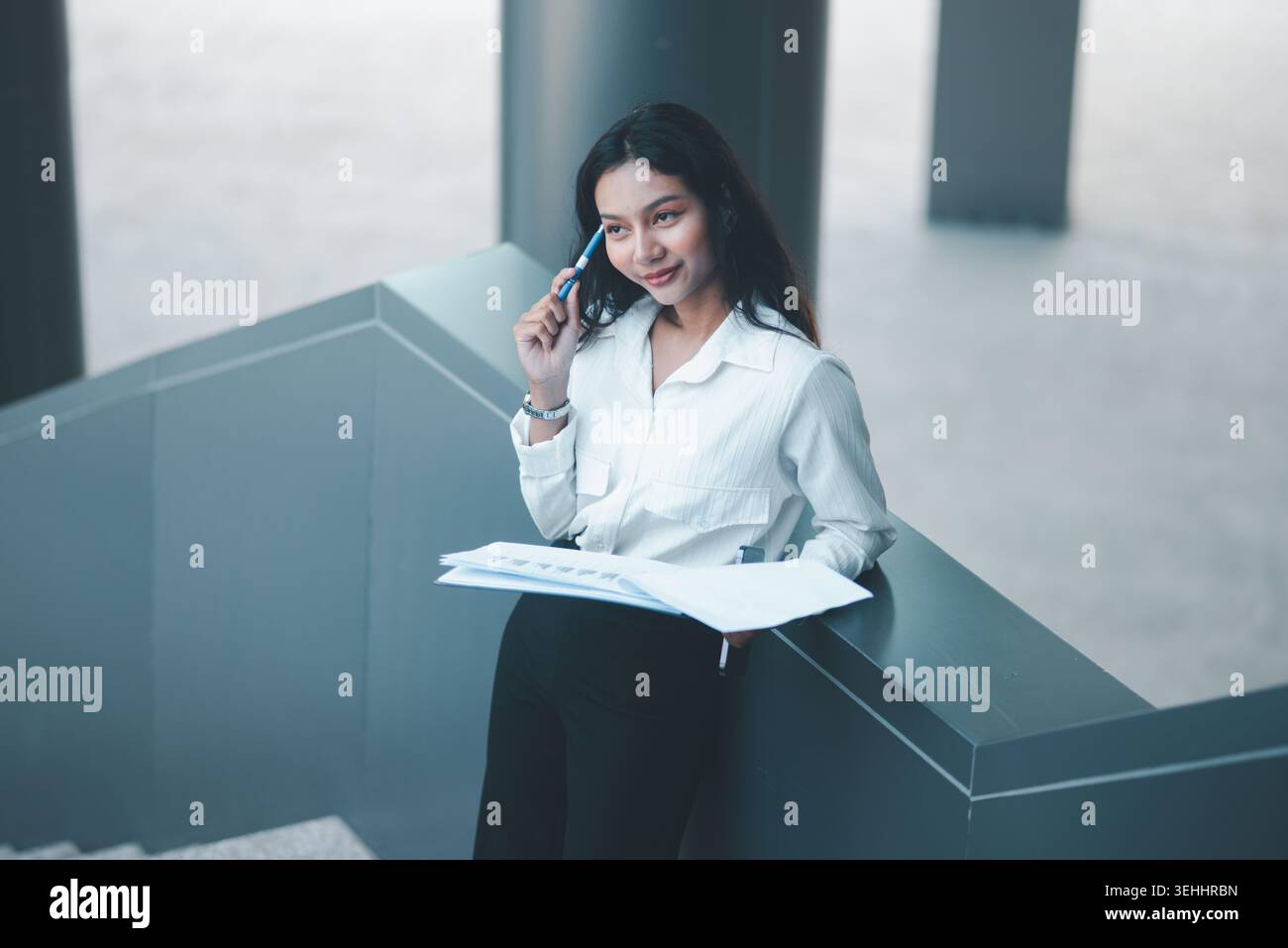 Donna d'affari professionista che tiene una penna al tempio mentre esamina documenti da una ringhiera di un ufficio, simboleggiando il pensiero strategico, la pianificazione e i dati Foto Stock