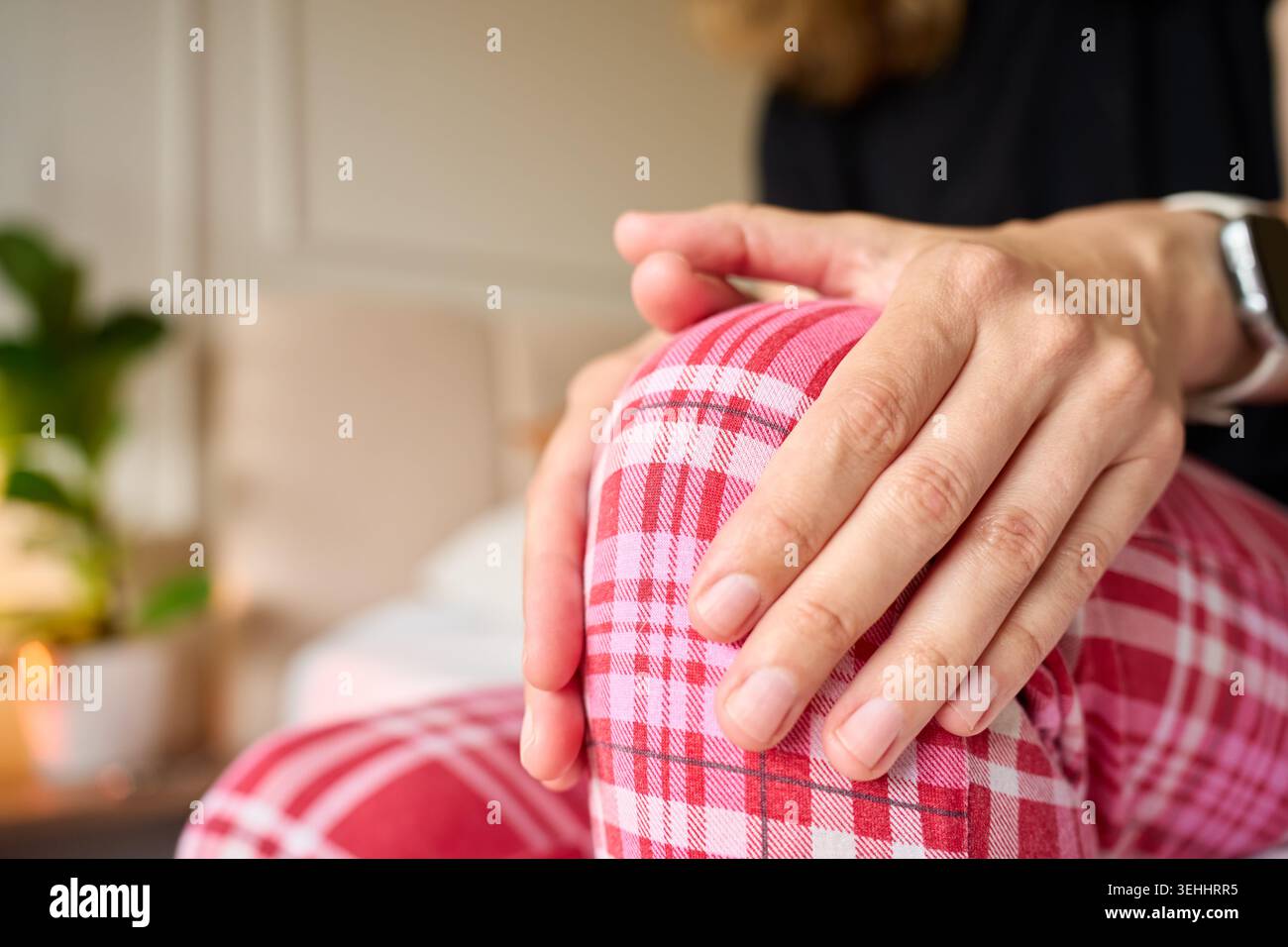 La donna soffre di dolore al ginocchio, massaggiando il ginocchio con entrambe le mani mentre è seduta sul letto. Concetto di mal di ginocchio, disagio articolare, problema di mobilità e casa Foto Stock