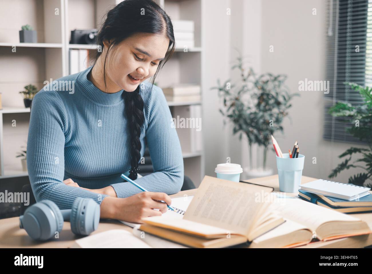 Giovane donna asiatica che scrive appunti di studio alla scrivania con libri aperti, caffè e cuffie nelle vicinanze. Autoapprendimento mirato, preparazione all'esame, lettura, istruzione Foto Stock