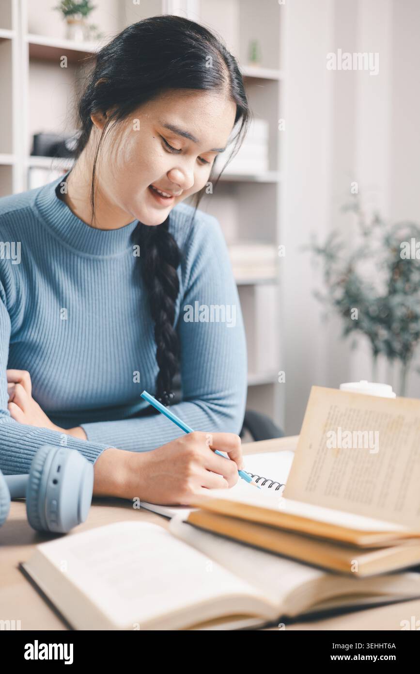 Giovane donna asiatica che scrive appunti di studio alla scrivania con libri aperti, caffè e cuffie nelle vicinanze. Autoapprendimento mirato, preparazione all'esame, lettura, istruzione Foto Stock