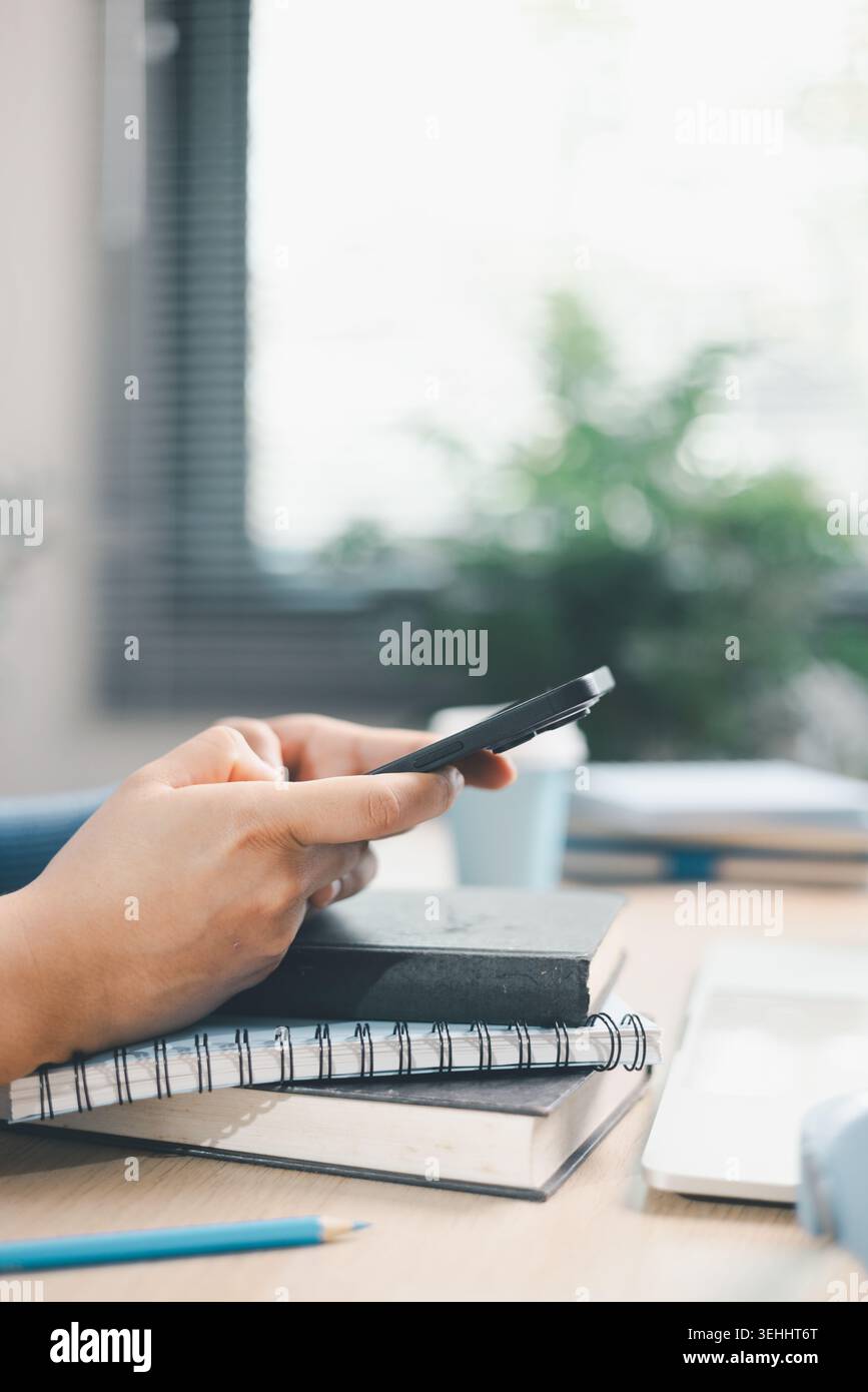Giovane donna che usa lo smartphone sopra i libri accanto al computer portatile e alle cuffie alla scrivania. Apprendimento mobile, studio online, ricerca digitale, multitasking, educ Foto Stock