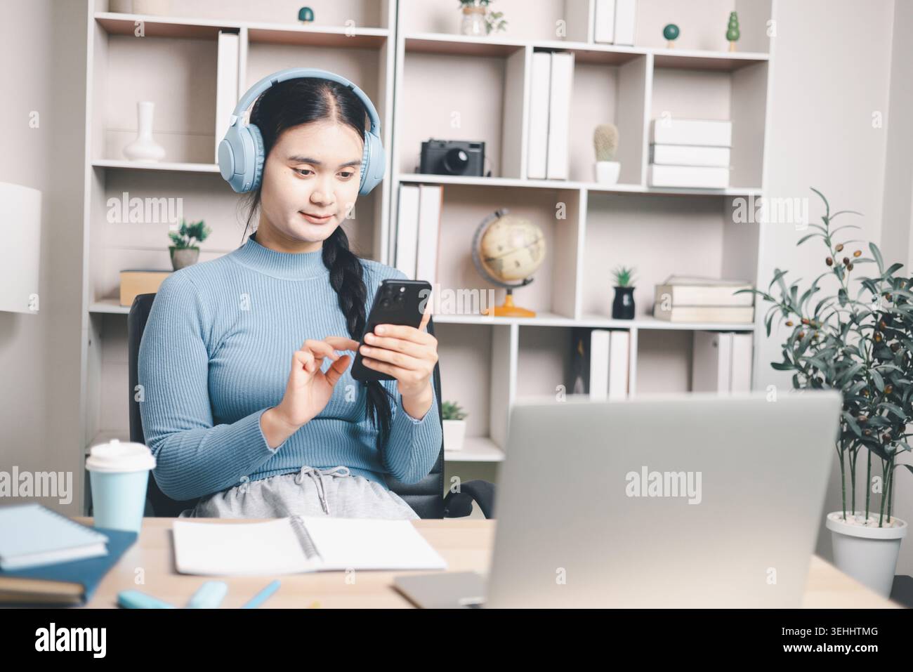 Giovane donna asiatica che indossa le cuffie e utilizza lo smartphone accanto al computer portatile alla scrivania. Apprendimento mobile, classe online, studio remoto, istruzione digitale A. Foto Stock