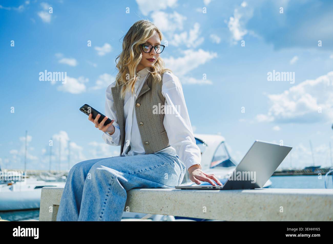 Una giovane donna siede su una panchina al porticciolo. Utilizza un computer portatile mentre tiene in mano un telefono. Le barche sono nell'acqua e il cielo è luminoso con le nuvole. Foto Stock