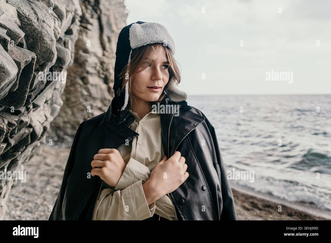 Donna ushanka Coast giovane donna che indossa una giacca di pelle e un cappello invernale si staglia sulla costa rocciosa del mare in un'atmosfera penosa all'aperto Foto Stock