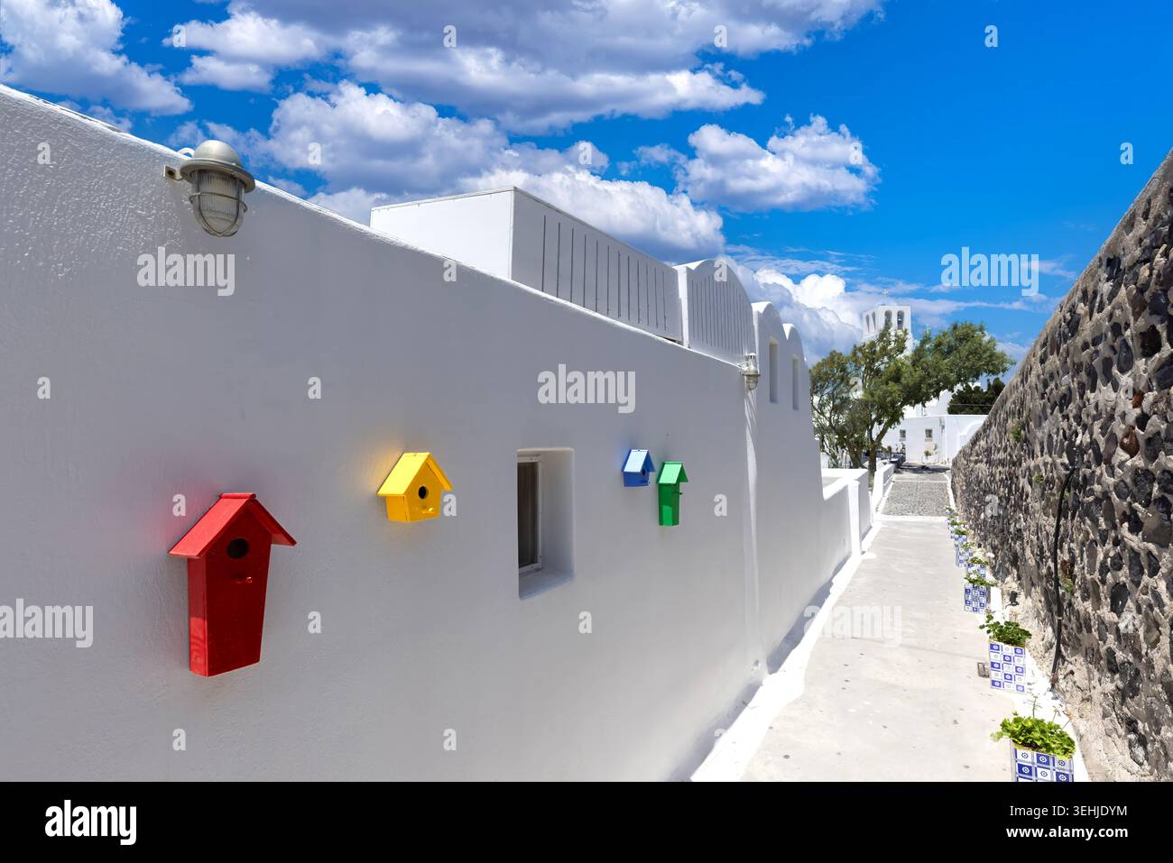 Vista delle colorate case per uccelli montate su un muro bianco lungo una stretta strada sotto un cielo blu con nuvole a Fira, Grecia. Foto Stock
