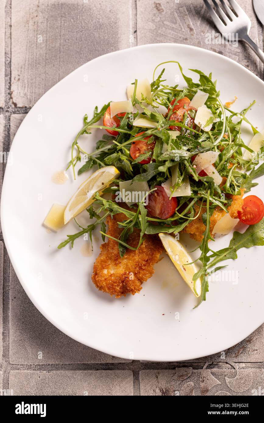 Un piatto appetitoso di filetto di pollo impanato servito con un contorno di rucola fresca, pomodori ciliegini, fette di parmigiano e spicchi di limone. Una t Foto Stock