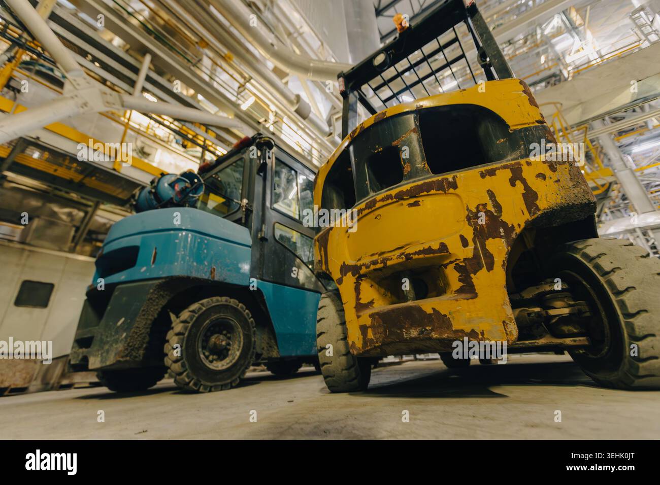 Due trattori industriali, uno blu e uno giallo usurato, si trovano all'interno di una grande struttura meccanica con travi metalliche, tubi e strutture sopraelevate. Foto Stock