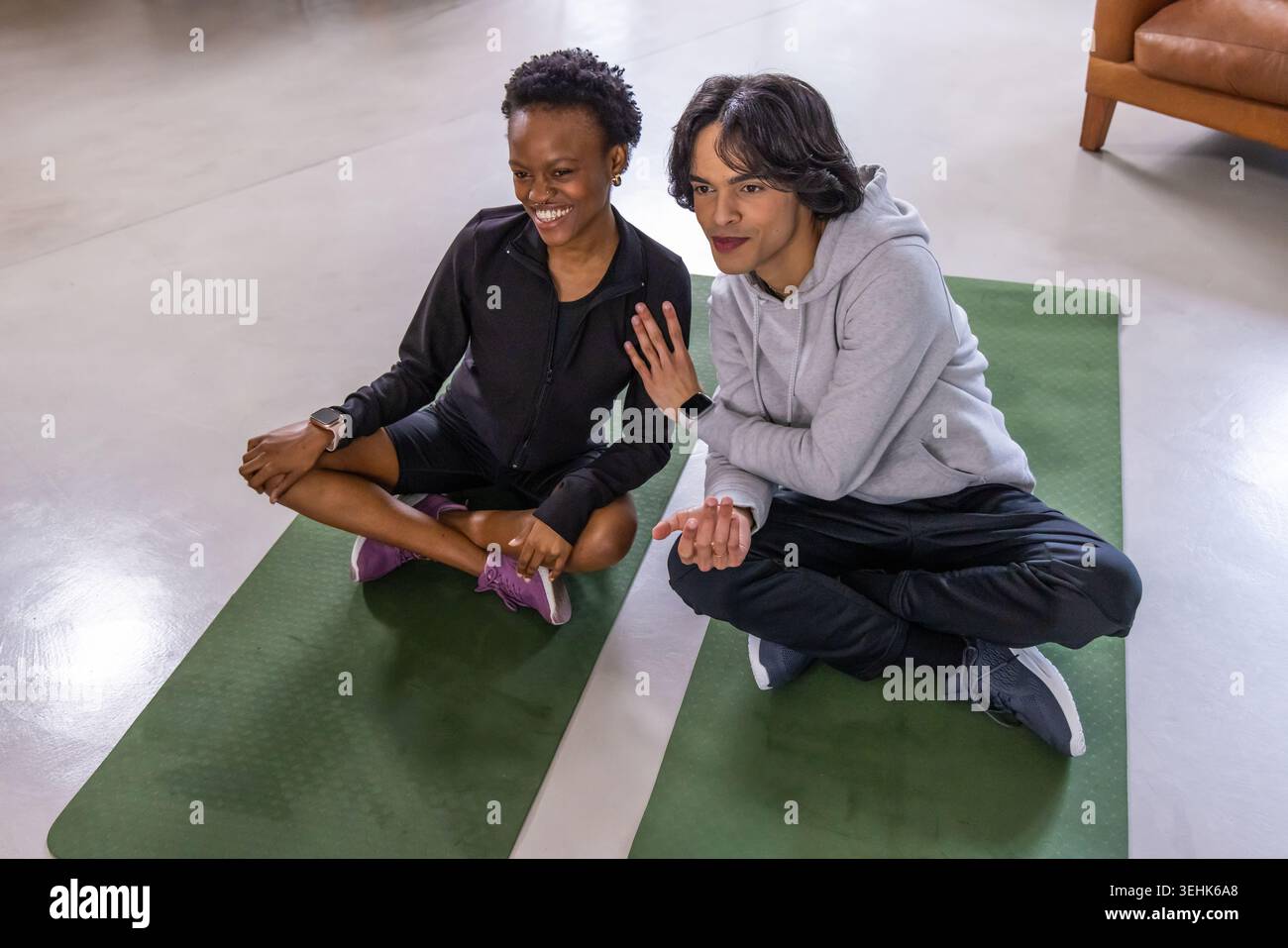 Amici diversi seduti su tappetini verdi in studio, chiacchierando, gestando, indossando abbigliamento da ginnastica e orologi Foto Stock