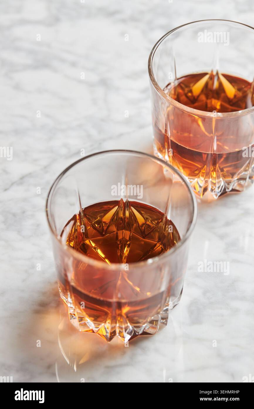 Primo piano di due bicchieri di cristallo riempiti di whisky bourbon color ambra su un piano di lavoro in marmo bianco, che mostrano colori ricchi, riflessi ed elegante bar st Foto Stock