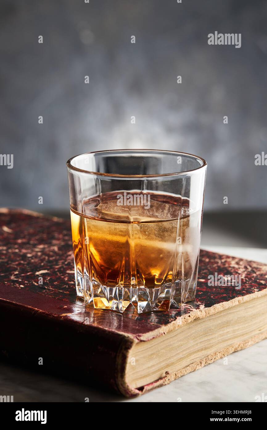 Primo piano del whisky bourbon in un bicchiere limpido su un vecchio libro consumato, bevanda calda color ambra su una superficie in marmo con luce soffusa e sfondo sfocato. Foto Stock