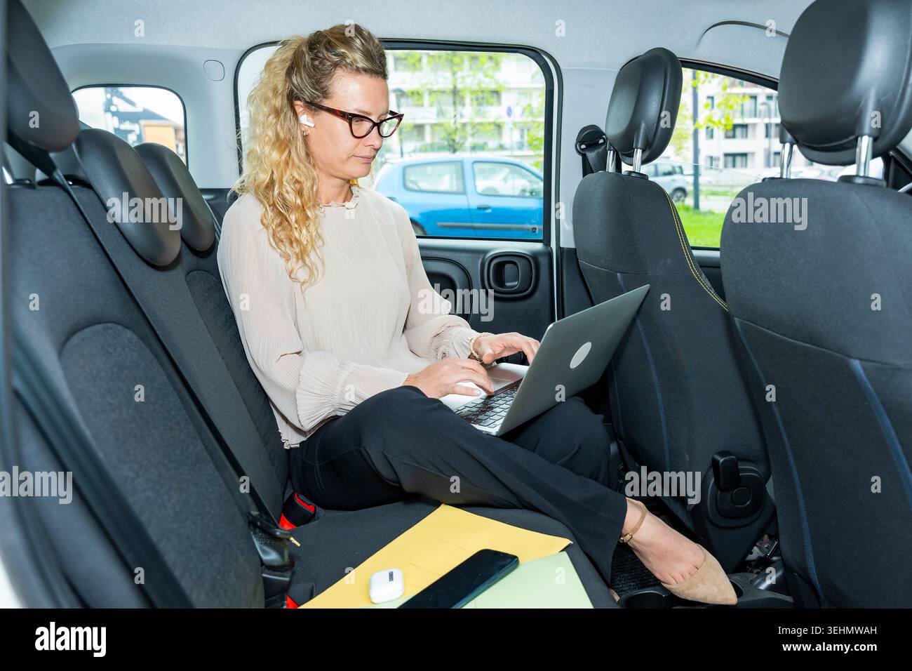 Una donna d'affari con occhiali lavora su un notebook sul sedile posteriore di un'auto, utilizzando auricolari e file wireless durante gli spostamenti in città, illustrando i dispositivi mobili o Foto Stock