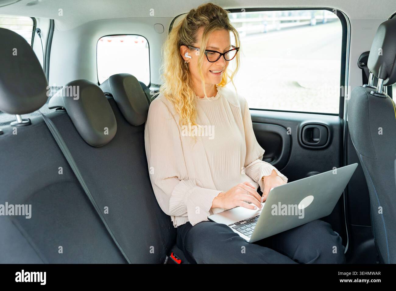 Una donna d'affari sorridente che indossa occhiali e auricolari lavora su un notebook sul sedile posteriore di un'auto, utilizzando il tempo di viaggio per lavorare in remoto, comunicare e. Foto Stock