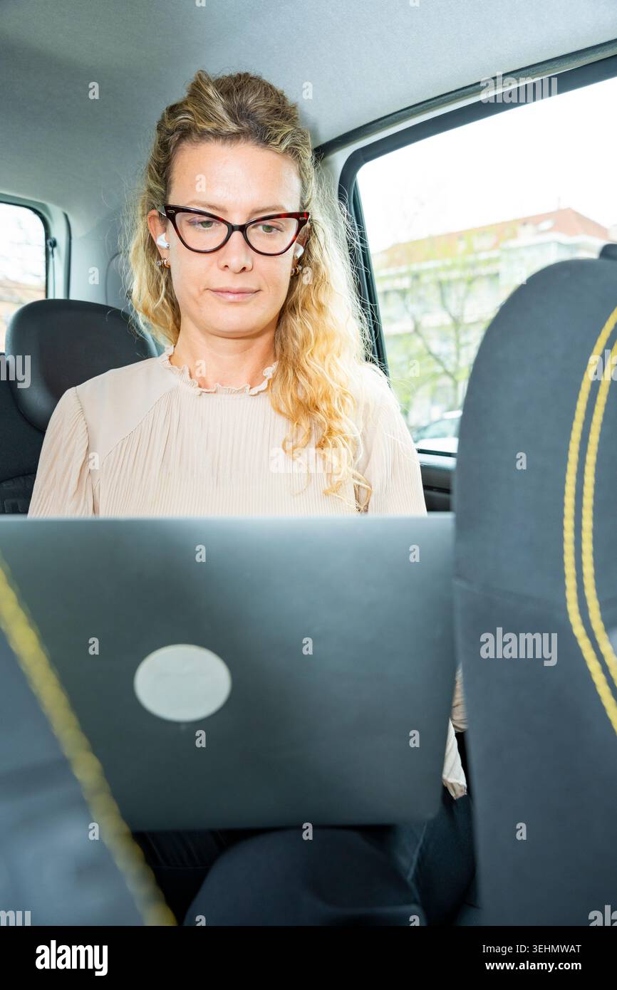 Una donna d'affari concentrata che indossa occhiali lavora su un notebook sul sedile posteriore di un'auto, utilizzando auricolari wireless durante gli spostamenti, che rappresentano l'offic mobile Foto Stock