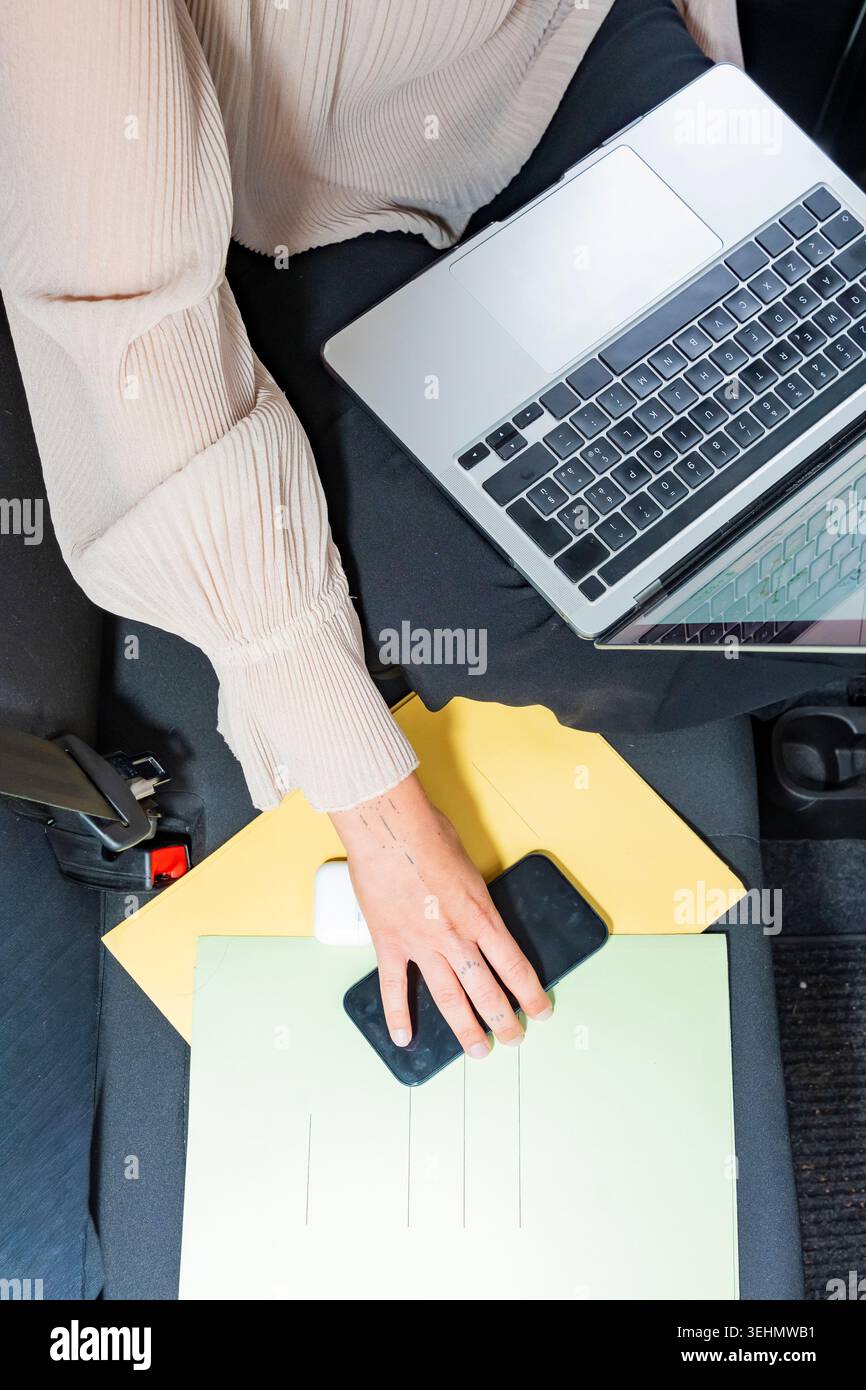 Vista dall'alto di una donna d'affari in un'auto che utilizza un laptop e uno smartphone con documenti e cartelle in grembo, gestendo attività e comunicazioni mentre trav Foto Stock