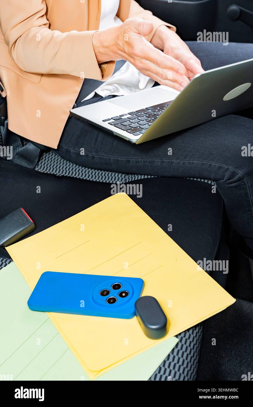 Primo piano di una donna d'affari che utilizza un laptop sul sedile del passeggero di un'auto, gestendo mentre lavora in remoto con documenti cartacei e uno smartphone durante un Foto Stock