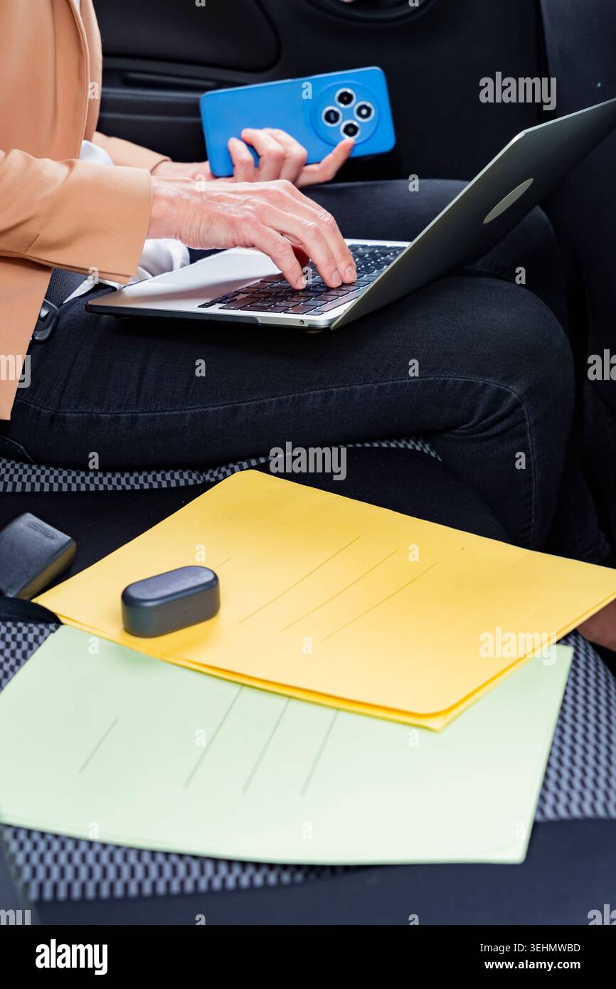Primo piano di una donna d'affari che lavora su un laptop e tiene in mano uno smartphone mentre è in macchina, con i documenti in grembo, che mostrano la produttività mobile Foto Stock