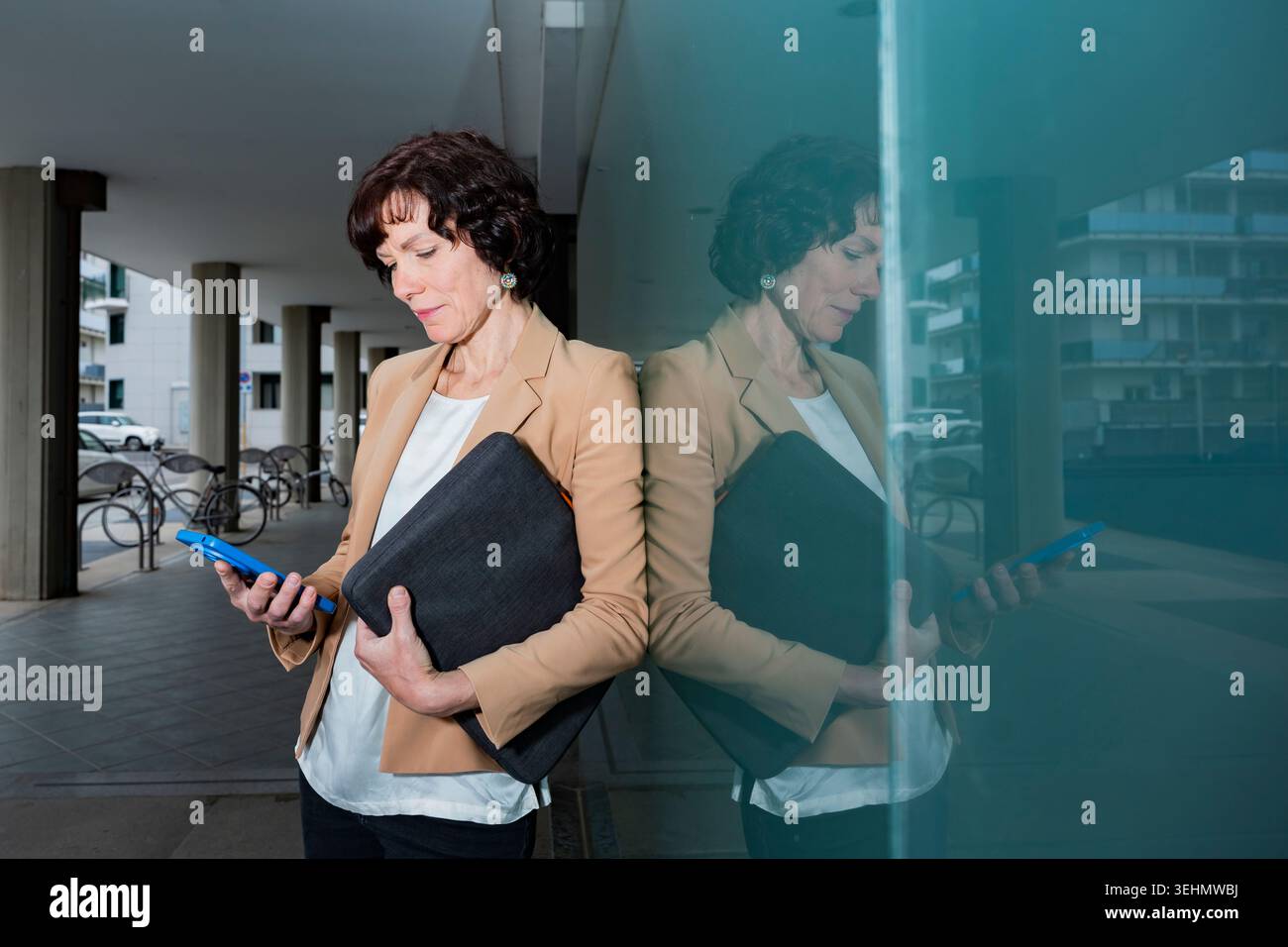 Una donna d'affari professionista si appoggia su una parete di vetro all'esterno di un moderno edificio per uffici, tenendo in mano una custodia per laptop e controllando uno smartphone, con un ripiano urbano Foto Stock