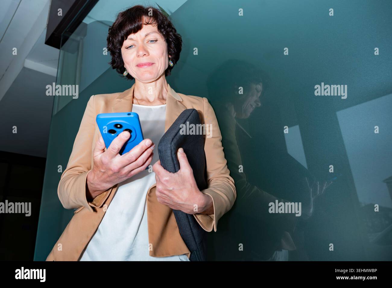 Una donna d'affari in blazer controlla uno smartphone mentre tiene in mano una custodia per laptop, accanto a una moderna parete di vetro. Stile di vita aziendale professionale, tecnologia Foto Stock