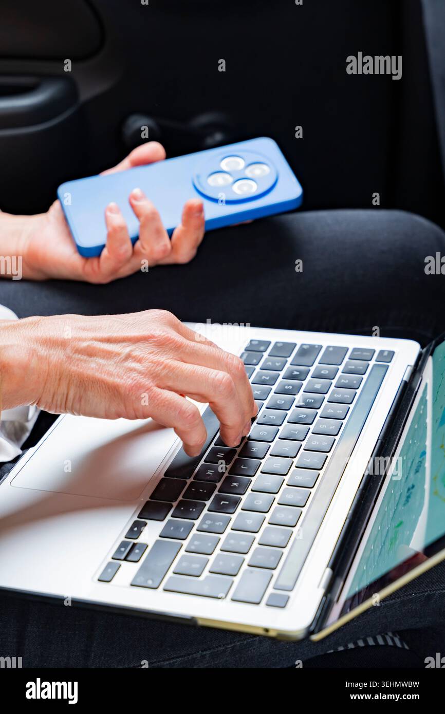 Primo piano di una donna d'affari che lavora sul sedile posteriore di un'auto, digita un laptop mentre tiene in mano uno smartphone, illustra la produttività mobile e i viaggi Foto Stock