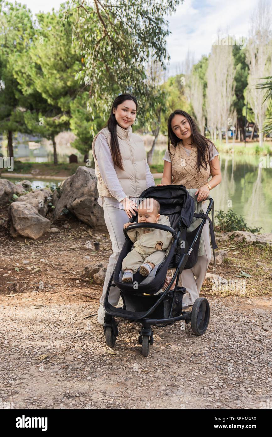Un momento di gioia in famiglia catturato in un tranquillo parco, con due donne che interagiscono felicemente con un bambino seduto in un passeggino, circondato dalla natura Foto Stock