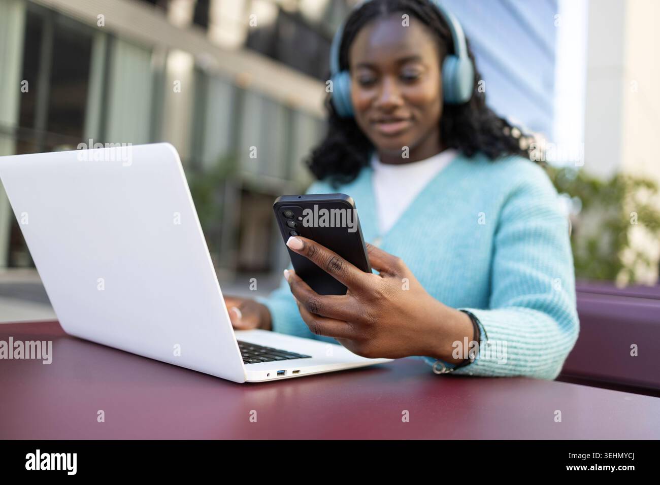 Una donna africana che indossa le cuffie lavora su un computer portatile mentre controlla il suo smartphone all'aperto in un moderno campus di uffici, mostrando lavoro remoto e tecnologia Foto Stock