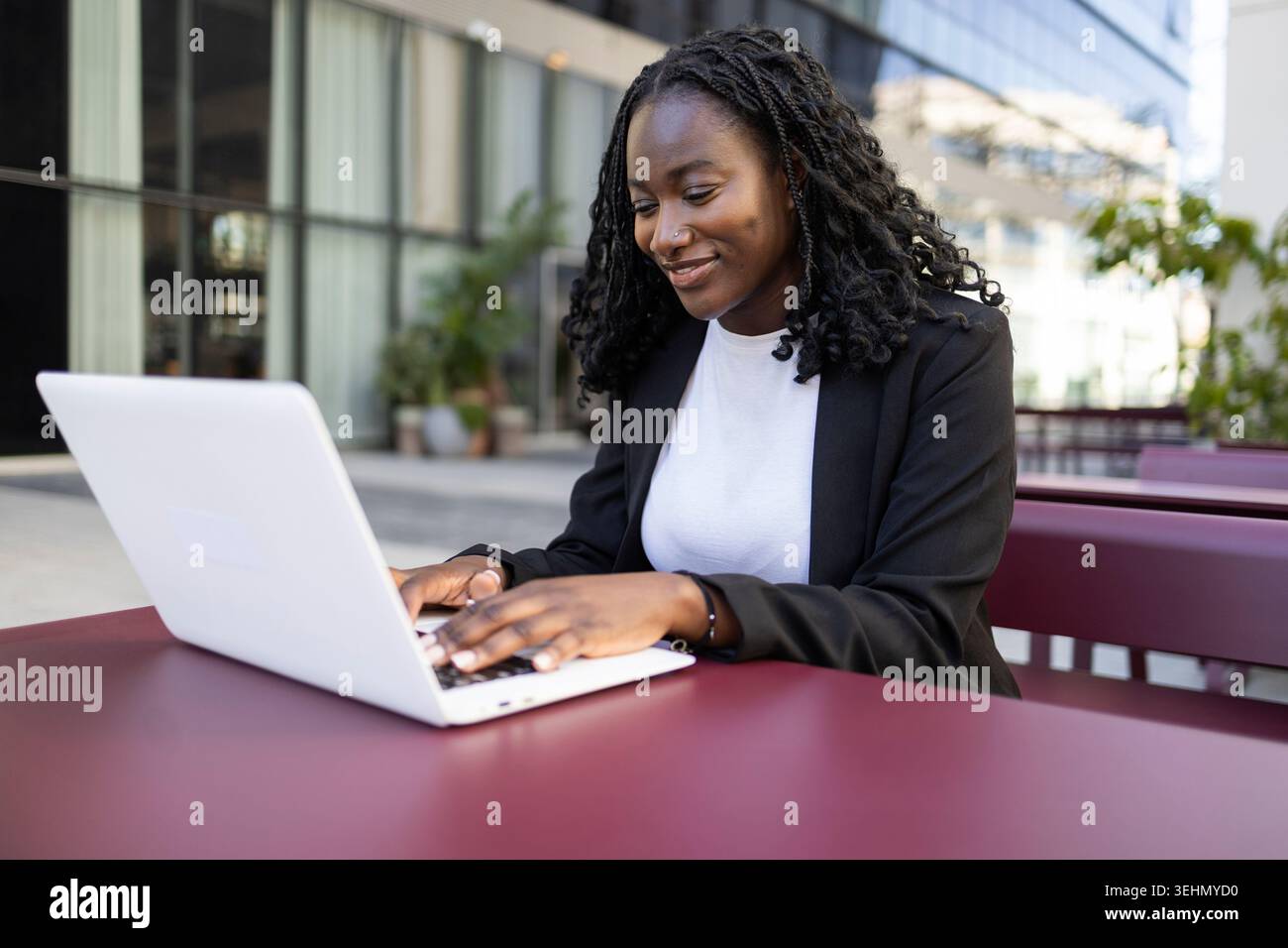 Donna nera africana sorridente in abiti da lavoro che lavora su un notebook a un tavolo all'aperto vicino a un moderno edificio di uffici, concentrata sul lavoro da remoto, lo studio, o Foto Stock