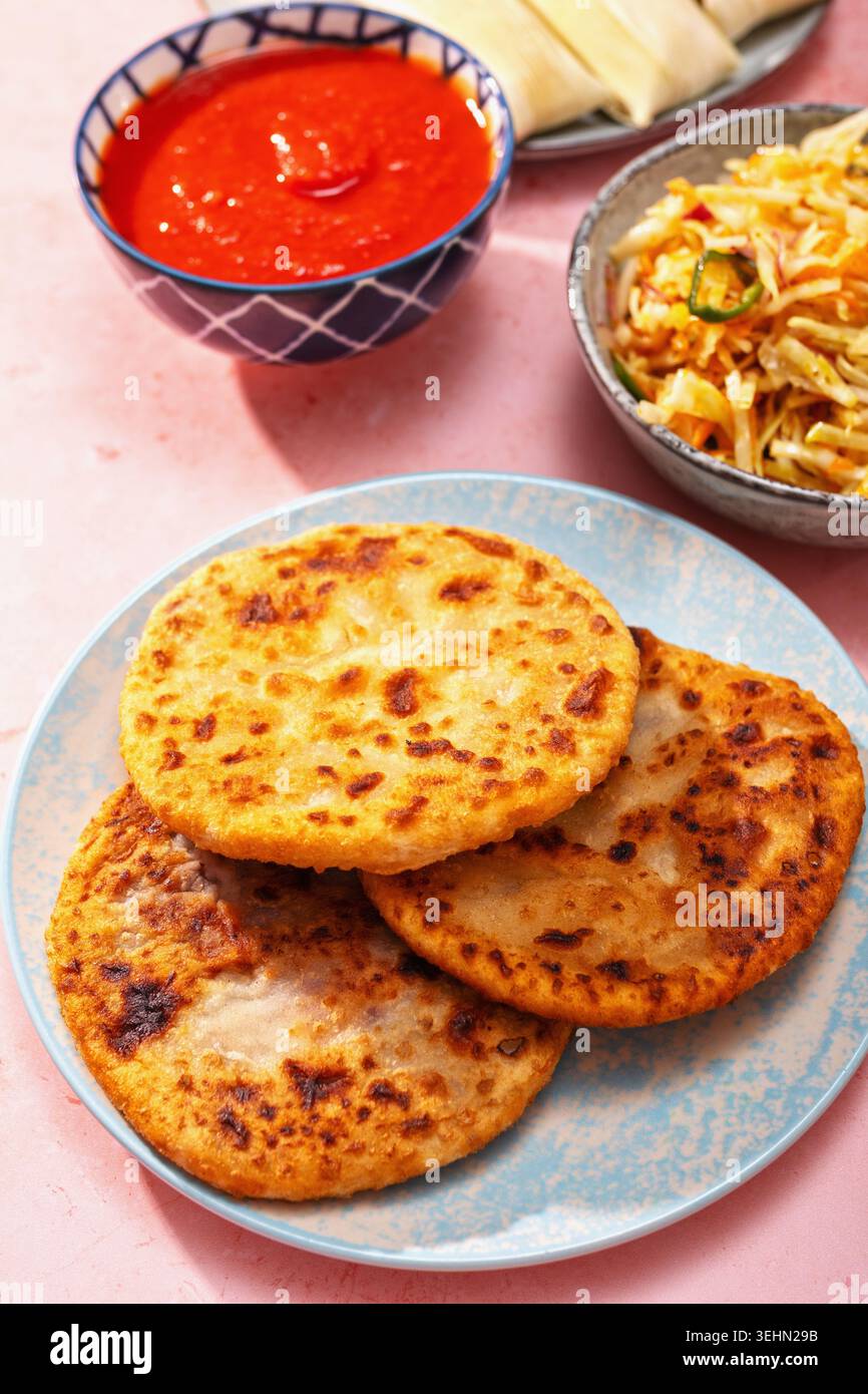 Vista dall'alto di piatti latino-americani con pupusas, arepas, curtido, humitas, yuca e salsa su un colorato tavolo rosa per una cena di famiglia. Foto Stock