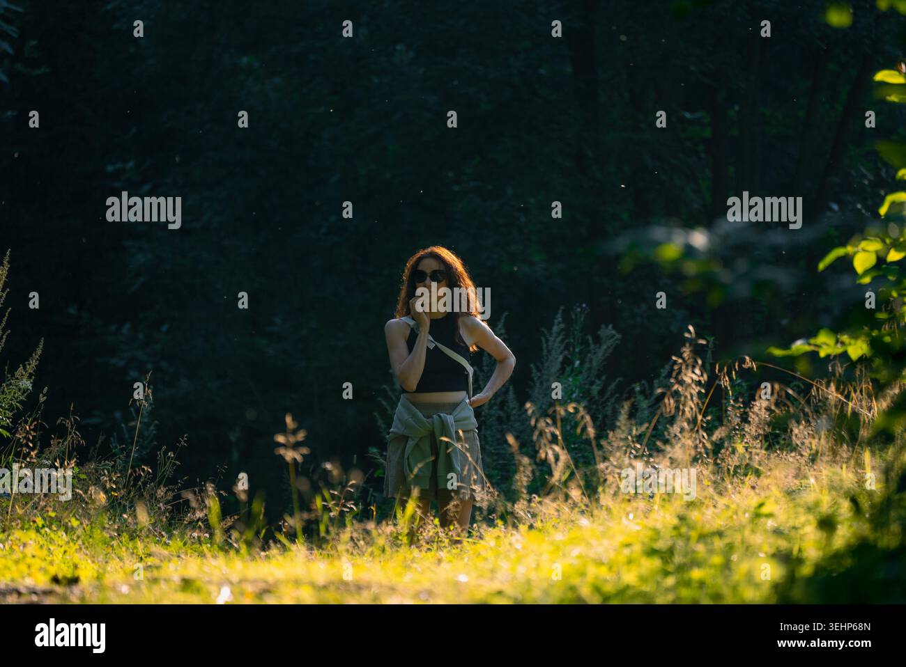 Una giovane donna che indossa gli occhiali da sole attraversa una tranquilla foresta al tramonto, assaporando la tranquillità della natura mentre si ferma per momenti di riflessione e contemplazione Foto Stock