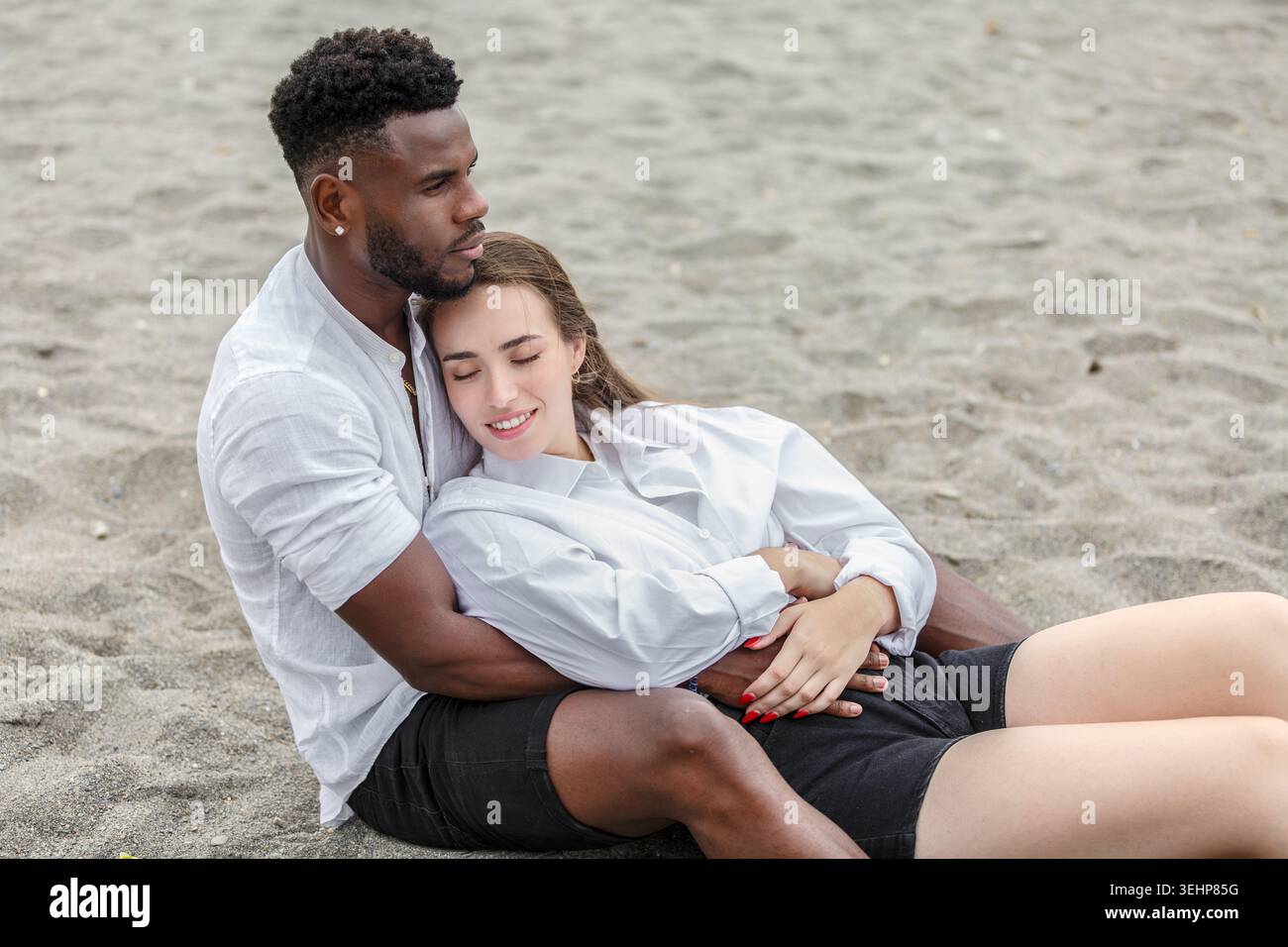 Una coppia si rilassa sulla spiaggia di sabbia, abbracciando delicatamente. L'uomo si siede con la schiena al sole, tenendo la donna vicina mentre si appoggia la testa sul suo sho Foto Stock
